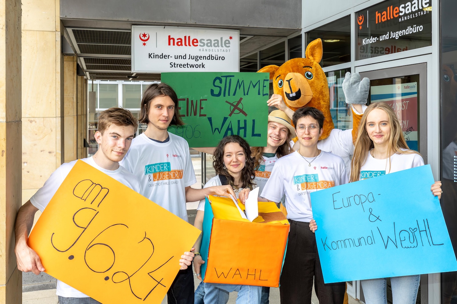 Sechs junge Menschen halten Plakate hoch mit dem Hinweis auf die Europa- und Kommunalwahl am 9. Juni 2024. Sie stehen vor dem Kinder- und Jugendbüro der Stadt Halle (Saale). Ein Känguru-Maskottchen in Lebensgröße steht hinter ihnen. Alle tragen ein T-Shirt des Kinder- und Jugendrates der Stadt Halle (Saale) und werfen Zettel in eine riesige Wahlurne.