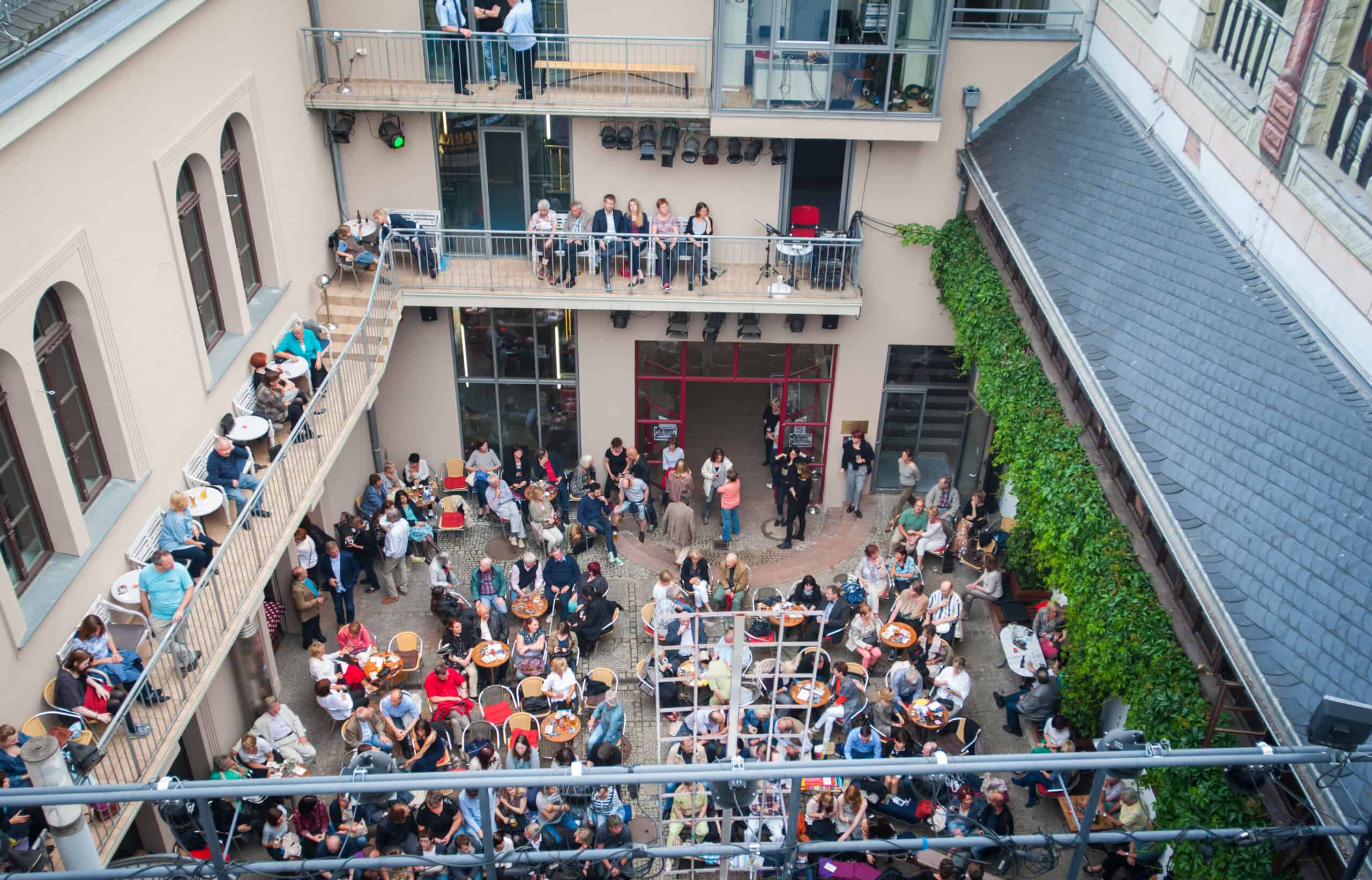Blick von oben auf eine gut besuchte Veranstaltung im Innenhof eines Gebäudes mit mehreren Balkonen. Menschen sitzen an runden Tischen, während andere stehen und sich unterhalten.