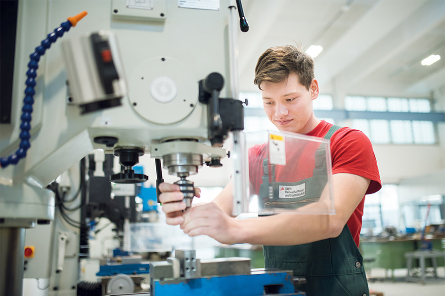 in junger Auszubildender in rotem T-Shirt und grüner Arbeitskleidung arbeitet konzentriert an einer großen Industriemaschine in einer modernen Werkstatt. Er hält ein Werkzeug in beiden Händen und fokussiert auf seine Arbeit, während er die Maschine bedient. Die Werkstatt ist hell und großzügig gestaltet, mit technischen Geräten und Arbeitsstationen im Hintergrund.