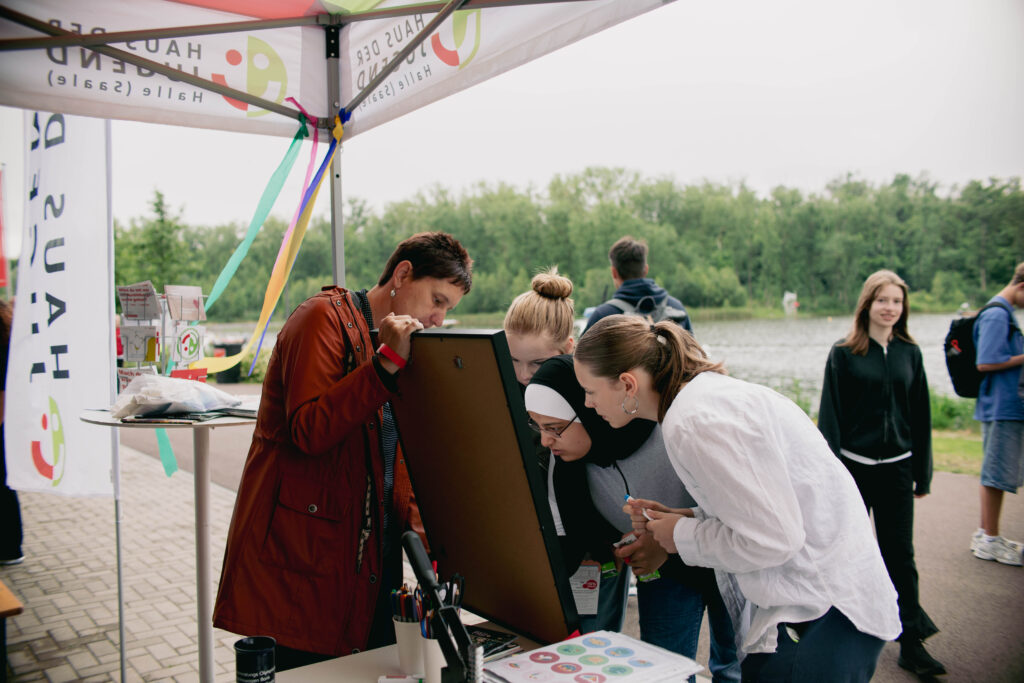 Vier Personen stehen dicht gedrängt um ein großes Wortpuzzle, das an einem Stand des Hauses der Jugend ausgestellt ist. Im Vordergrund ist eine Person in einer roten Jacke zu sehen, die sich konzentriert über das Rätsel beugt. Drei junge Frauen, eine davon mit Kopftuch, betrachten ebenfalls interessiert das Puzzle. Im Hintergrund sind ein See und weitere Personen zu erkennen. Der Stand ist mit bunten Bändern und dem Logo des Hauses der Jugend dekoriert.