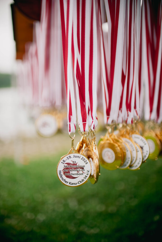 Nahaufnahme von Medaillen mit rot-weißen Bändern, die beim 23. Sparkassencup der Schulen im Drachenboot am 18.-19. Juni 2024 vergeben wurden. Die Medaillen tragen die Aufschrift "23. Sparkassencup der Schulen im Drachenboot" und "Hallescher Kanu-Club 54 e.V." und hängen in einer Reihe bereit für die Gewinner*innen. Im Hintergrund sind unscharf weitere Medaillen und grüne Wiese sowie der Osendorfer See zu erkennen.