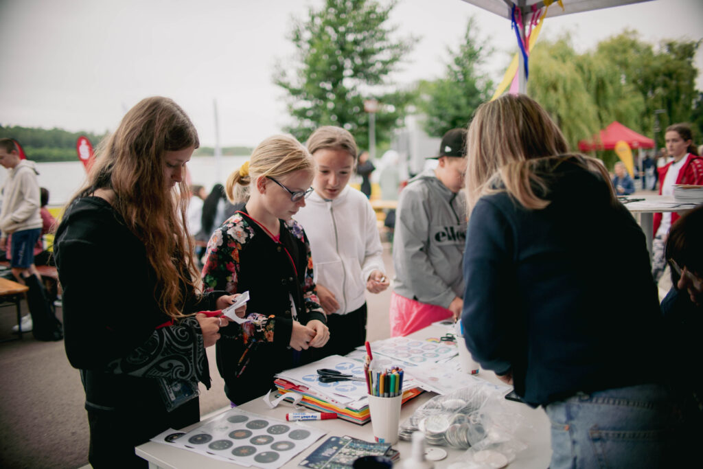 Mehrere Kinder und Jugendliche stehen um einen Tisch an einem Stand des Hauses der Jugend. Sie sind beschäftigt mit dem Gestalten von Buttons und benutzen dafür Scheren, Papier und bunte Stifte. Der Tisch ist mit verschiedenen Materialien und Vorlagen bedeckt. Im Hintergrund sind weitere Teilnehmer*innen und Stände zu sehen, die eine lebhafte und aktive Atmosphäre vermitteln. Der See und grüne Bäume sind ebenfalls im Hintergrund erkennbar.