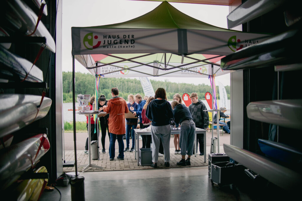 Blick von innen auf einen Stand des Hauses der Jugend, der sich direkt vor einem Gebäude befindet. Der Stand ist mit einem großen Pavillon überdacht und bunt dekoriert. Mehrere Personen, darunter Jugendliche und Erwachsene, stehen um Tische herum und sind in verschiedene Aktivitäten vertieft. Im Hintergrund sind ein See, grüne Bäume und weitere Veranstaltungselemente wie Flaggen und Stände zu erkennen. Links im Bild sind die Rümpfe von Drachenbooten oder Kanus im Gebäudeinneren zu sehen.