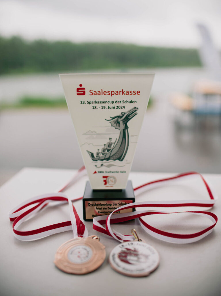 Eine Nahaufnahme von einem Pokal und zwei Medaillen, die auf einem Tisch liegen. Der Pokal trägt die Aufschrift "Saalesparkasse 23. Sparkassencup der Schulen 18. - 19. Juni 2024" und zeigt ein Bild von einem Drachenboot. Die Medaillen sind an rot-weißen Bändern befestigt. Im Hintergrund ist unscharf die grüne Umgebung des Sees zu erkennen.