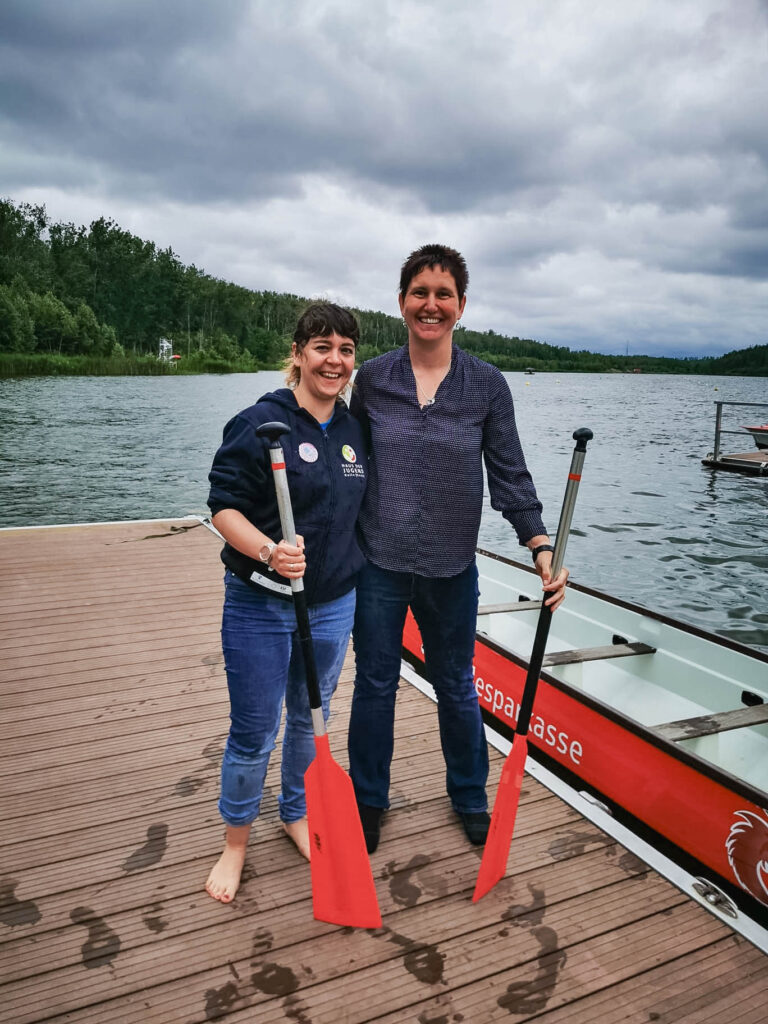 Zwei Frauen stehen lachend auf einem Steg am See, jeweils mit einem Paddel in der Hand. Die Frau links trägt eine blaue Jeans und ein dunkelblaues Kapuzenpullover, während die Frau rechts ein blaues Hemd und dunkle Hosen trägt. Im Hintergrund sind ein rotes Drachenboot und der bewölkte Himmel zu sehen.