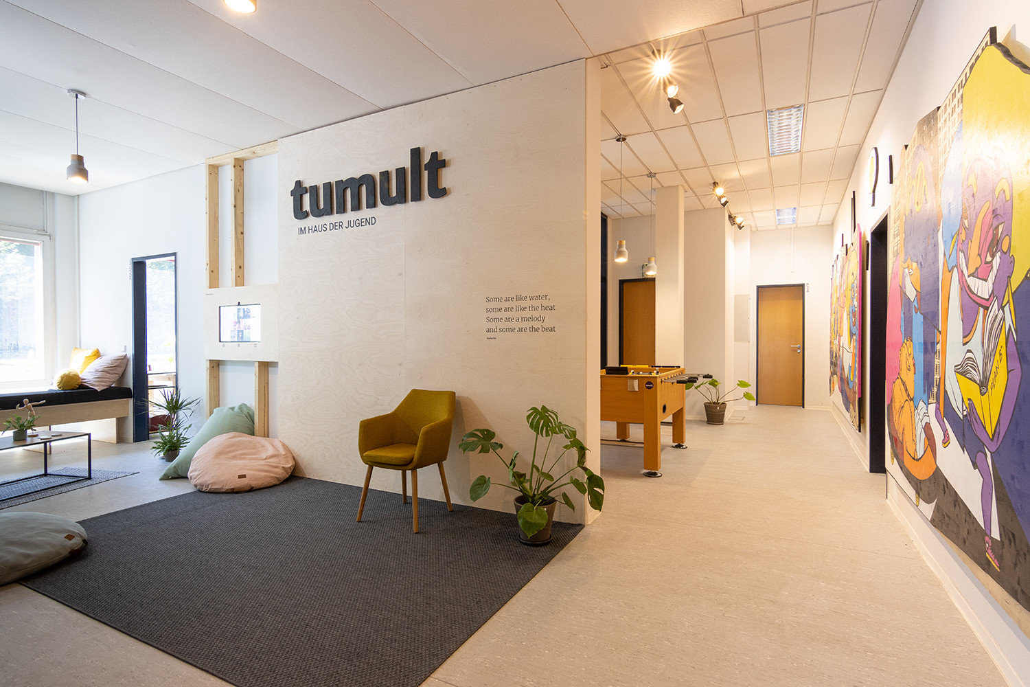 Ein einladender Innenraum der Jugendberatungsstelle tumult im Haus der Jugend in Halle (Saale). Die moderne Einrichtung umfasst eine gemütliche Sitzecke mit Sitzkissen, einen Kicker-Tisch sowie bunte Wandgemälde. Die warme Beleuchtung und grünen Pflanzen schaffen eine entspannte Atmosphäre. An der Wand prangt das tumult-Logo mit dem Zusatz „IM HAUS DER JUGEND“.