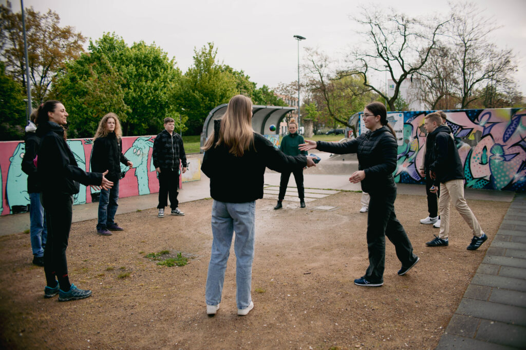 Junge Menschen stehen im Kreis vor einem Skateplatz und werfen sich Bälle bei einem Kennenlernspiel zu.