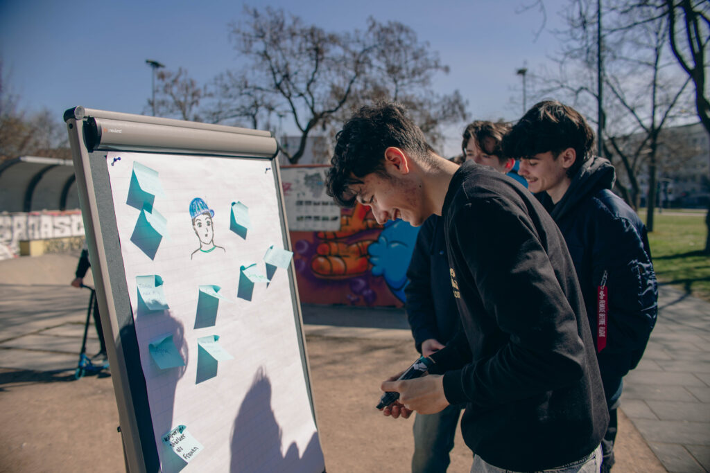 Draußen bei Sonnenschein kleben Jugendliche blaue Zettel an ein Flipchart mit einer gezeichneten Figur.
