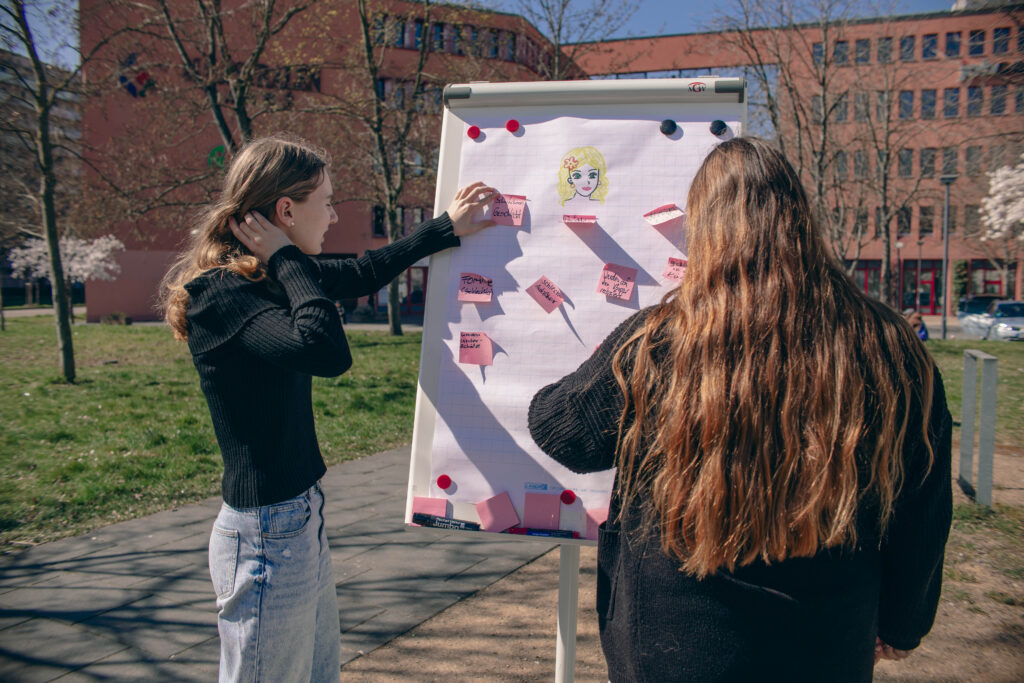 Zwei Mädchen stehen draußen vor einem Flipchart mit einer Zeichnung und pinken Notizzetteln. Sie diskutieren über die Inhalte.