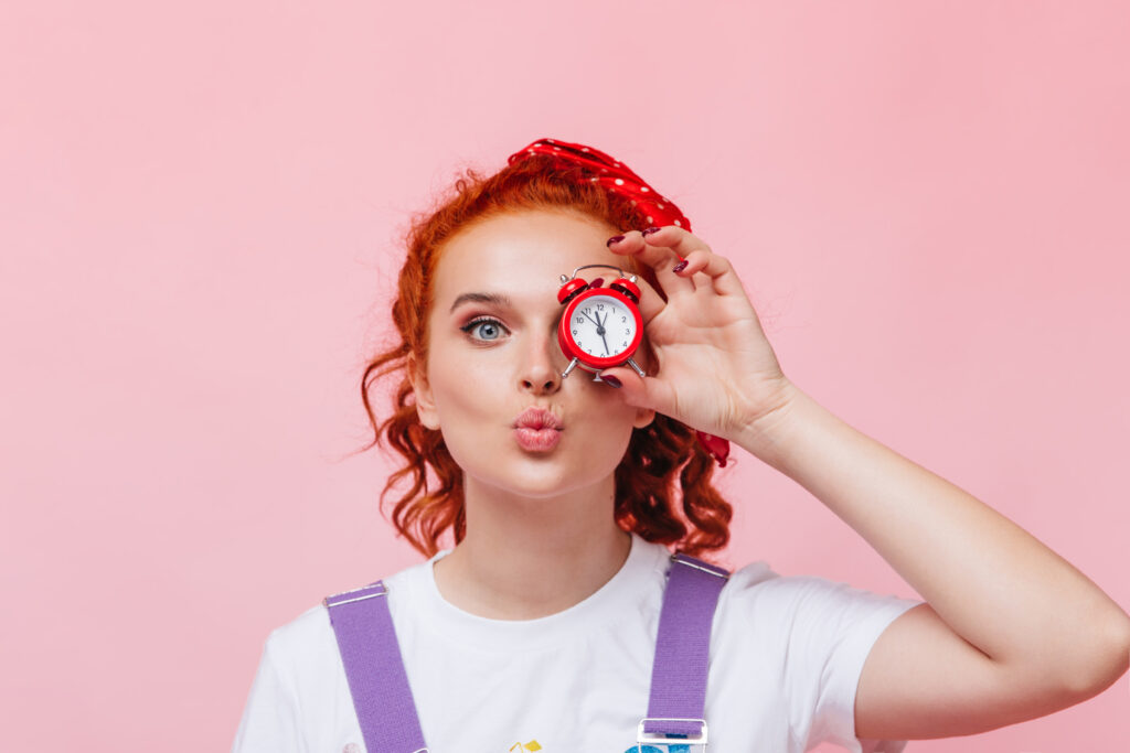Perky girl in white T-shirt blows kiss and covers eye with alarm clock