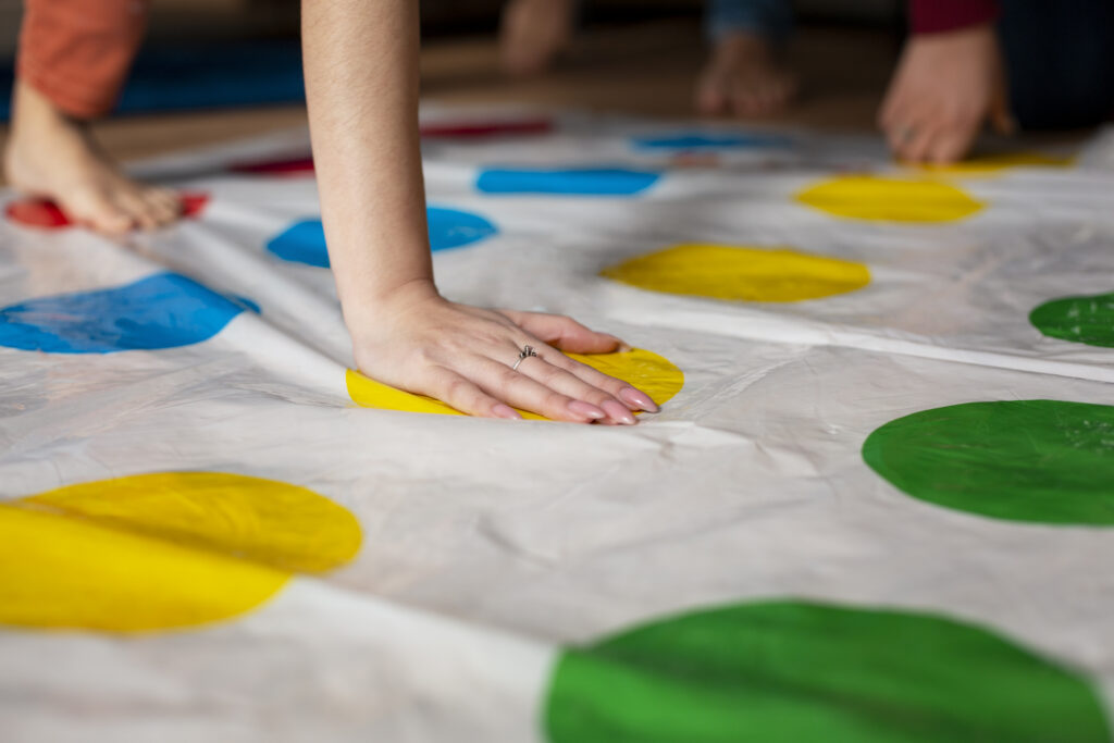 Mehrere Personen spielen gemeinsam Twister; im Vordergrund ist eine Hand auf einem gelben Kreis zu sehen.