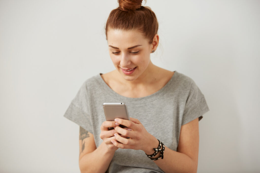 Portrait of a smiling young woman using cell phone. Beautiful happy girl chatting and texting on mobile phone on the grey background. Young woman reading a text message on smart phone